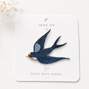 May include: An iron-on embroidered patch of a swallow in flight, featuring dark blue, light blue, white, and orange-red thread. The patch is mounted on a white card labelled IRON ON and little dove studio with a small dove icon and heart symbols.