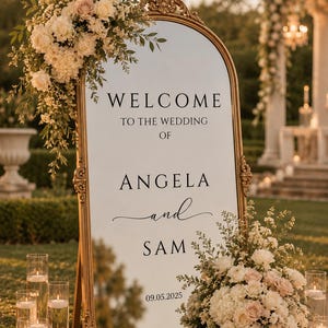 May include: A gold-framed mirror with the words "WELCOME TO THE WEDDING OF ANGELA and SAM" displayed. The mirror is adorned with floral arrangements and candles, with the date 09.05.2025 at the bottom. The scene is set outdoors.