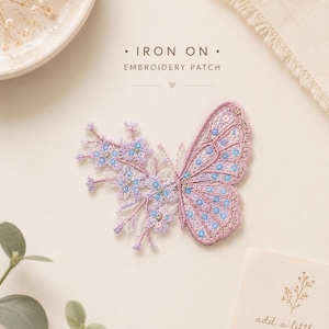 Puede incluir: Un parche bordado termoadhesivo con forma de mariposa, donde una ala está compuesta por delicadas ramitas florales en azul y violeta claro, y la otra ala presenta intrincados patrones de hilo en rosa y azul. El texto dice IRON ON EMBROIDERY PATCH.