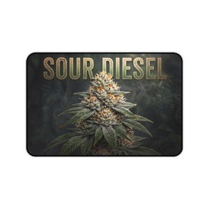 May include: A rectangular desk mat featuring a central cannabis flower bud with green leaves and orange pistils. The text SOUR DIESEL is printed in a large, bold, metallic-toned font across the top against a dark, smoky background.
