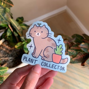 May include: A sticker featuring a brown cat with a sprout on its head, sitting next to a potted plant. The sticker has a light blue background with the words "PLANT COLLECTOR" in white text. Two small star shapes are on the sticker.