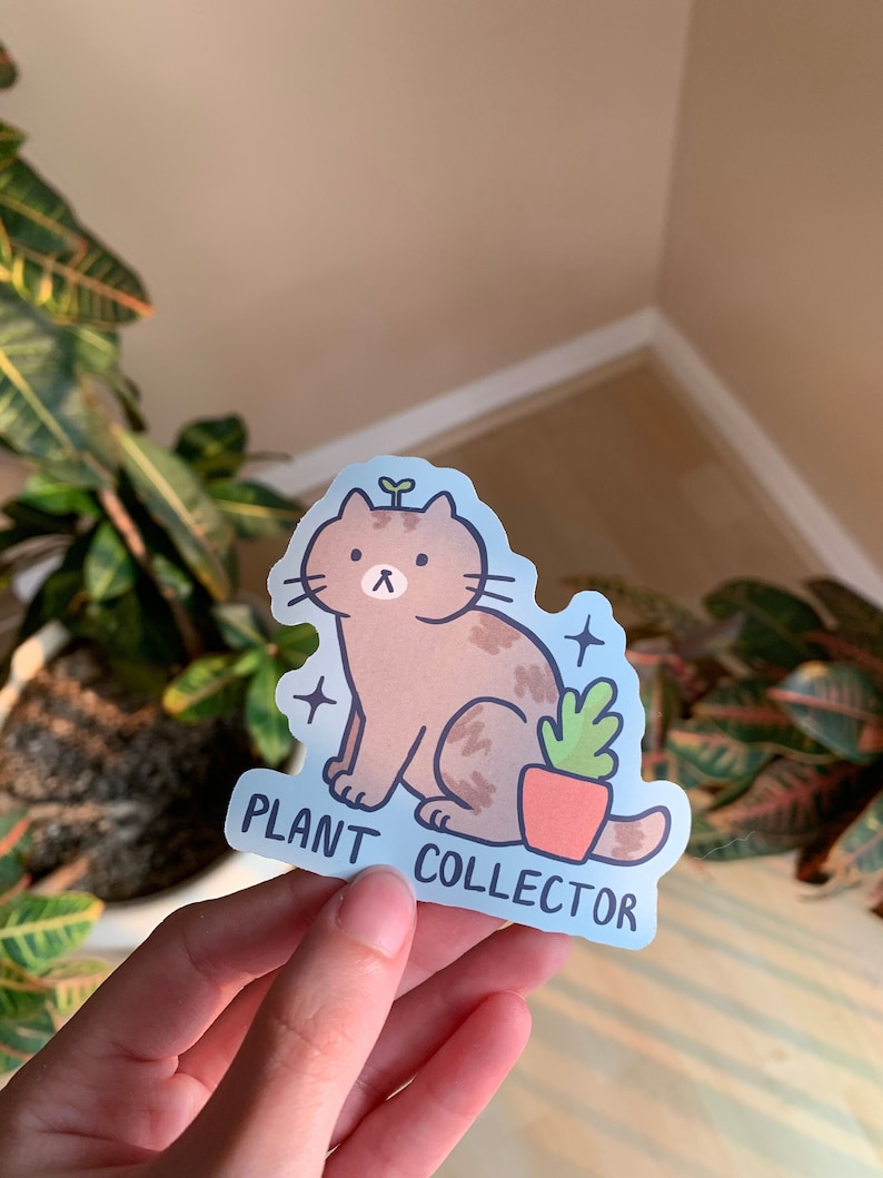 May include: A sticker featuring a brown cat with a sprout on its head, sitting next to a potted plant. The sticker has a light blue background with the words "PLANT COLLECTOR" in white text. Two small star shapes are on the sticker.