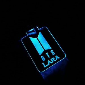 Può includere: Un portachiavi LED blu con il logo BTS e il nome "LARA" scritto sotto.