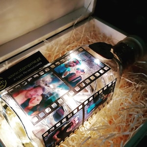 May include: A glass bottle with a cork top, decorated with photos and string lights. The bottle is in a white box with wood shavings. The bottle has a business card attached to it with the text "www.stampme.pt Artigos Personalizados".