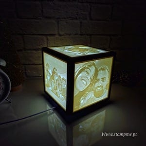以下が含まれることがあります： 各面に写真が彫られた、光る木製のキューブランプ。 ランプは黒いフレームで、暖かい黄色の光を発し、画像を表示します。 ウェブサイトのアドレス「www.stampme.pt」が表示されています。