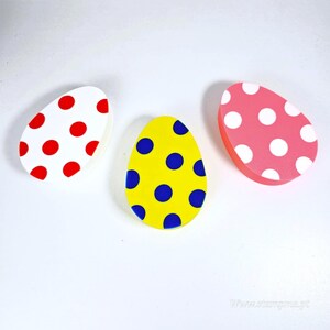 Può includere: Tre oggetti decorativi a forma di uovo di Pasqua. Uno è bianco con pois rossi, un altro è giallo con pois blu e il terzo è rosa con pois bianchi. Le uova sono disposte su una superficie bianca.