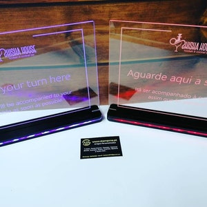 May include: Two acrylic signs with LED lights, one with the text "Wait your turn here. You will be accompanied to your table as soon as possible." and the other with the text "Aguarde aqui a sua vez. Irá ser acompanhado à assim que." Both signs have the logo "Shisha House" in the top left corner.