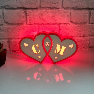 Op de afbeelding: Twee in elkaar grijpende houten harten met de letters C en M in het midden, met een ampersand ertussen. De harten worden verlicht met rood licht, waardoor een schaduw op het witte oppervlak eronder valt.