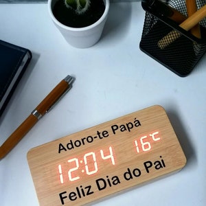 Puede incluir: Un reloj de madera con una pantalla digital roja que muestra la hora 12:04 y la temperatura de 16 grados Celsius. El reloj tiene el texto "Adoro-te Papá" y "Feliz Dia do Pai" grabado.