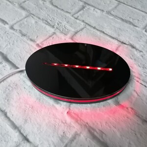 Peut inclure: Un présentoir rotatif rond noir avec une bande de lumière LED rouge autour du bord. Le présentoir est sur une surface en brique blanche.