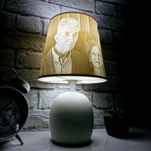 Peut inclure: Une lampe de table blanche avec un abat-jour crème orné d'une photo en noir et blanc d'un couple âgé. L'abat-jour est à rayures.