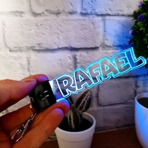 Könnte beinhalten: Ein transparenter Acryl-Schlüsselanhänger mit dem Namen "RAFAEL" in fetten, weißen Buchstaben. Der Schlüsselanhänger wird mit blauem Licht beleuchtet und hat einen schwarzen Metallring.
