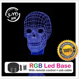 Puede incluir: Una lámpara con forma de calavera LED azul con un diseño de rejilla. La lámpara se encuentra sobre una base negra e incluye un mando a distancia y un cable USB. Se muestra el texto "RGB Led Base".