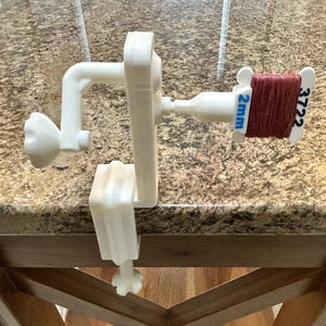 Könnte beinhalten: Ein weißer, 3D-gedruckter Fadenaufwickler, der an einer Oberfläche befestigt ist und eine Spule braunen Fadens hält. Auf der Spule steht "2mm" und "3722". Der Aufwickler hat eine Kurbel und eine Klemme.