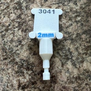 Può includere: Uno strumento di plastica bianca con il numero "3041" stampato in alto. Sotto, la misura "2mm" in blu. Lo strumento ha una forma unica con un corpo cilindrico e una piccola punta stretta.