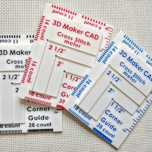 May include: Three white plastic cross-stitch tools with red and blue markings. Each tool has measurements in inches, including 2 1/2", 2", and 1 1/2". The tools are labeled "3D Maker CAD Cross Stitch meter" and "Corner Guide".