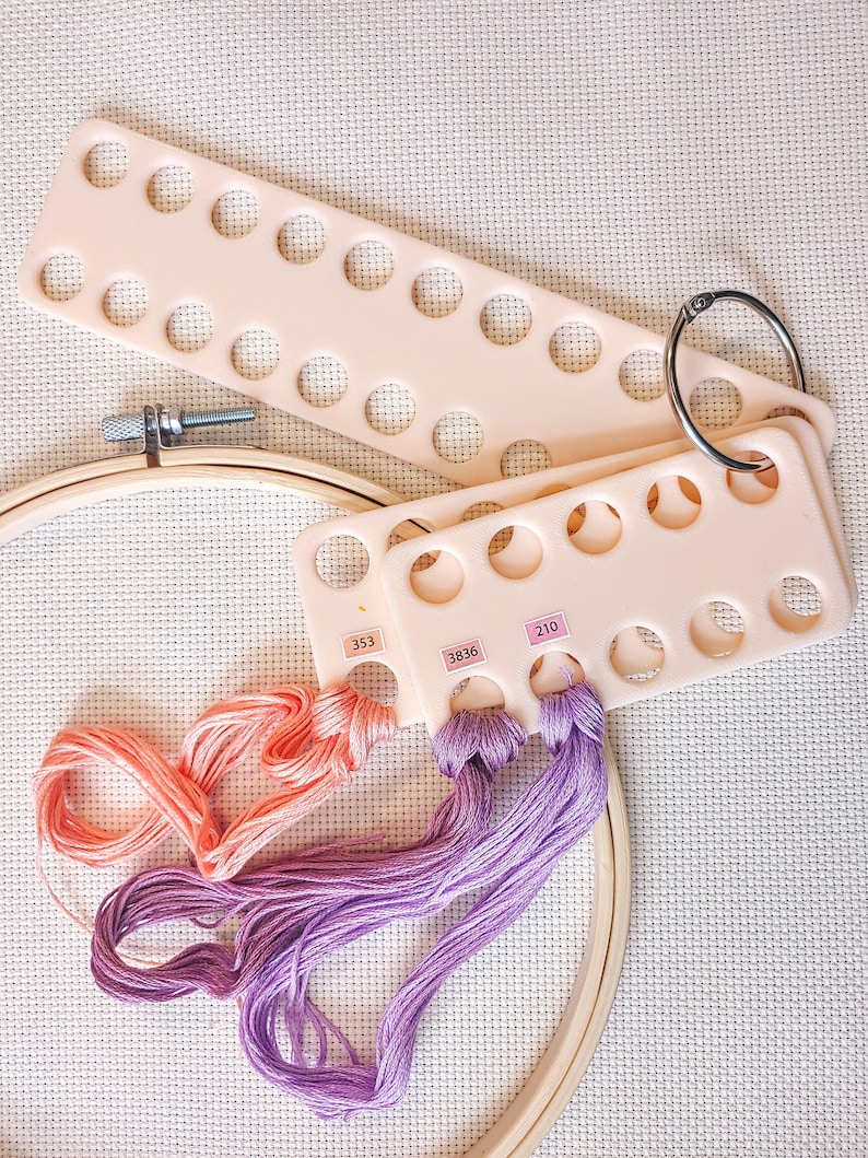May include: A close-up of a pink plastic thread organizer with three rows of holes. The organizer has a metal ring attached to it. There are three skeins of embroidery floss in shades of pink and purple, with the numbers 353, 3836, and 210 printed on the organizer.