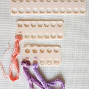 May include: Three white plastic thread organizers with rows of circular holes. The organizers are stacked on top of each other. Three skeins of embroidery floss are visible, one orange, one purple, and one lavender. The floss is threaded through the holes in the organizers. The numbers 353, 3836, and 210 are visible on the organizers.