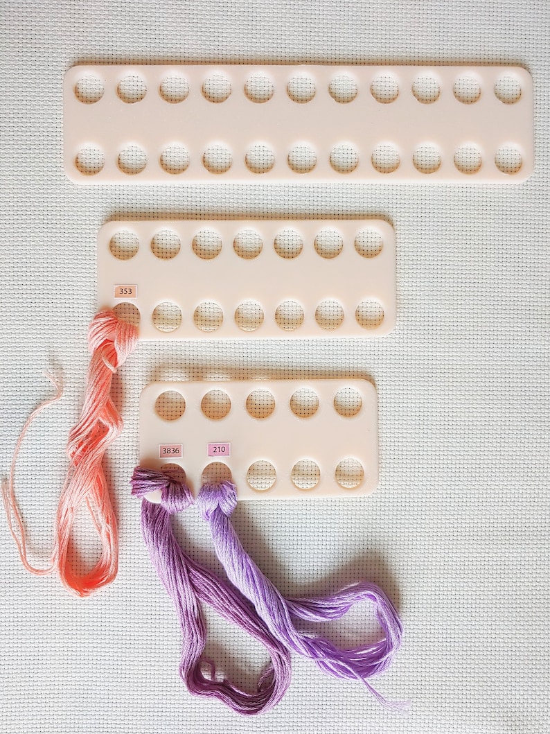 May include: Three white plastic thread organizers with rows of circular holes. The organizers are stacked on top of each other. Three skeins of embroidery floss are visible, one orange, one purple, and one lavender. The floss is threaded through the holes in the organizers. The numbers 353, 3836, and 210 are visible on the organizers.