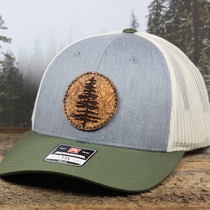 May include: A gray and green trucker hat with a brown leather patch featuring a tree design. The patch is stitched onto the front of the hat.