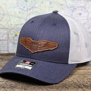 May include: A navy blue and white trucker hat with a brown leather patch featuring a winged American shield. The hat is made by Richardson 115.