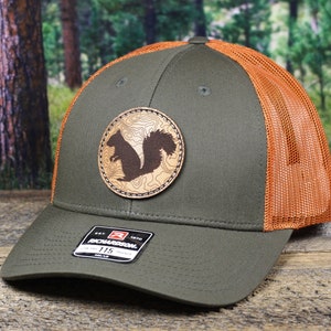 May include: A green and orange trucker hat with a brown leather patch featuring a silhouette of a squirrel. The patch has a topographic design.