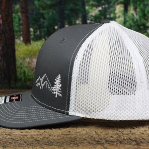 May include: A gray and white trucker hat with a white mesh back. The front of the hat features an embroidered design of mountains and a pine tree.