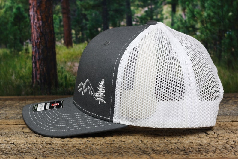 May include: A gray and white trucker hat with a white mesh back. The front of the hat features an embroidered design of mountains and a pine tree.
