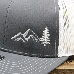 May include: A gray and white trucker hat with a white embroidered design of mountains and a pine tree. The hat has a mesh back and a curved brim.