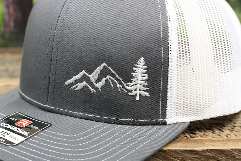 May include: A gray and white trucker hat with a white embroidered design of mountains and a pine tree. The hat has a mesh back and a curved brim.