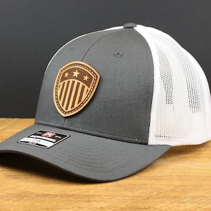 May include: A gray and white trucker hat with a wooden patch featuring a shield design with stars and stripes. The hat has a curved brim and a mesh back. The hat is displayed on a wooden surface.