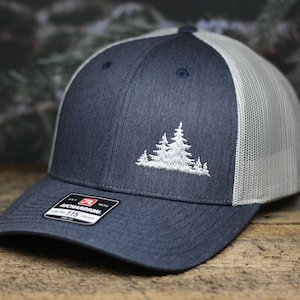 May include: A navy blue and white trucker hat with a white embroidered design of a group of trees. The hat has a mesh back and a Richardson 112 label.
