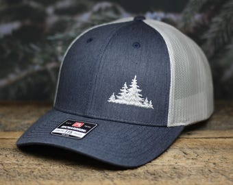 Può includere: Un cappello da camionista blu navy e bianco con un design ricamato bianco di un gruppo di alberi. Il cappello ha una parte posteriore in rete e un'etichetta Richardson 112.