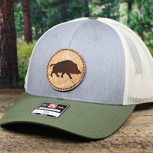 May include: A gray and green trucker hat with a brown leather patch featuring a wild boar silhouette. The patch has a topographic design around the boar.