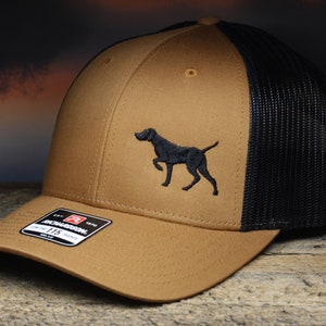May include: A brown and black trucker hat with a black embroidered German Shorthaired Pointer silhouette on the front. The hat has a black mesh back and a brown front panel. The hat has a Richardson 112 label on the side.