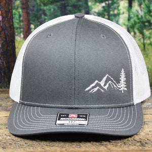 May include: A gray and white trucker hat with a white embroidered mountain and tree design. The hat has a mesh back and a curved brim.