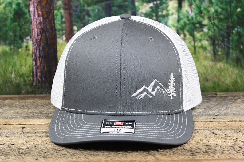 May include: A gray and white trucker hat with a white embroidered mountain and tree design. The hat has a mesh back and a curved brim.