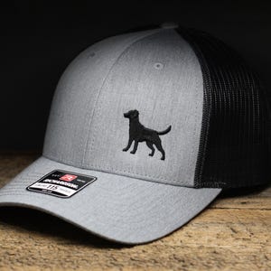 May include: A gray and black trucker hat with a black embroidered silhouette of a dog on the front. The hat has a structured front panel and a mesh back. The brim is curved, and the hat has an adjustable snap closure.