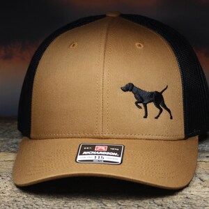 May include: A brown and black trucker hat with a black embroidered image of a dog on the front. The hat has a black mesh back and a brown front panel. The hat has a Richardson brand label on the front.