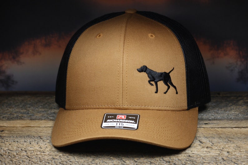 May include: A brown and black trucker hat with a black embroidered image of a dog on the front. The hat has a black mesh back and a brown front panel. The hat has a Richardson brand label on the front.
