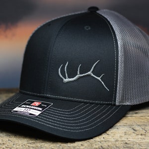 May include: Black and gray trucker hat with a gray embroidered antler design. The hat features a curved brim, a mesh back, and a snapback closure. The Richardson 112 hat has a structured fit and a pre-curved visor.