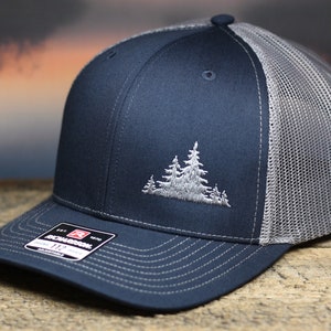 May include: A navy blue and gray trucker hat with a white embroidered design of three pine trees. The hat has a mesh back and a curved brim.