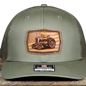 May include: Olive green trucker hat with a mesh back and a curved brim. The front features a leather patch with a tractor design over an American flag. The hat has a snapback closure and a small tag that reads "RICHARDSON 112".