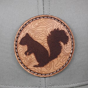 May include: A gray baseball cap featuring a circular patch with a brown squirrel silhouette. The patch has a topographic map design in a lighter brown color, with white stitching around the edge.