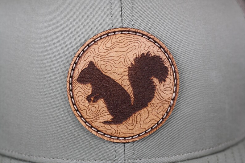 May include: A gray baseball cap featuring a circular patch with a brown squirrel silhouette. The patch has a topographic map design in a lighter brown color, with white stitching around the edge.