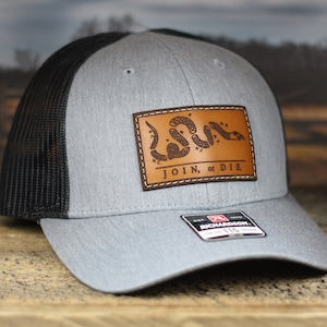 May include: Gray and black trucker hat with a leather patch. The patch features a coiled snake and the words "JOIN, or DIE". The hat has a mesh back and a curved brim.