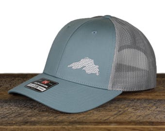 May include: A light blue baseball cap with a gray mesh back. The cap features a white embroidered outline of a land mass on the front. The brim is curved, and a black tag is visible on the side.