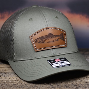 May include: A green mesh trucker hat with a brown leather patch featuring a trout design. The patch has white stitching around the edges. The hat has a black and red logo that says "Richardson 115" on the front.