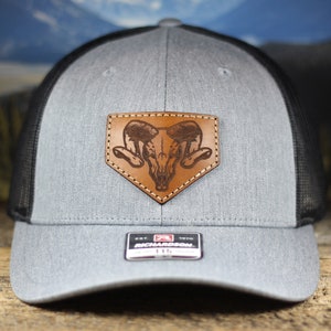 May include: A gray and black trucker hat with a brown leather patch featuring a skull with ram horns. The patch is stitched onto the front of the hat.