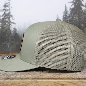May include: A sage green baseball cap with a mesh back and a curved brim. The cap features a rectangular patch with a design and a small label on the brim. The hat is displayed on a wooden surface.